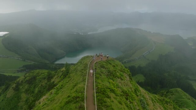 Aerial perspective of hikers on the narrow ridge trail at Boca do Inferno, overlooking the vast Sete Cidades volcanic caldera and its scenic lakes, partially obscured by dramatic, shifting mountain