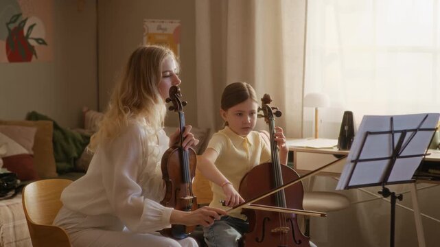 Medium long shot of Caucasian young adult teacher playing violin and instructing female child playing cello during home lesson. Ideal for music school marketing and educational campaigns