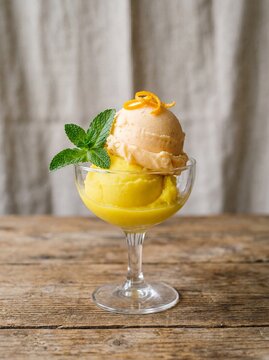 A professional photography shot of a double scoop of Limone and Mandarino ice cream in a glass dish, garnished with a fresh mint leaf and orange zest, professional photography, neutral background.
