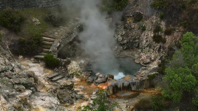 Aerial close-up of a geothermal fumarole in Furnas, Sao Miguel Island, featuring a steaming volcanic vent surrounded by mineral-stained rocks and stone steps built into the hillside.