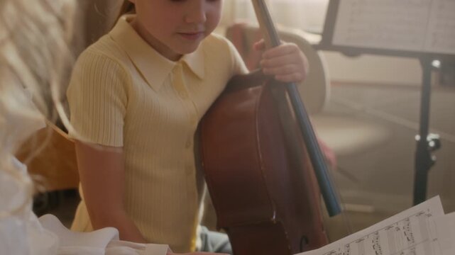 Tilt up shot of female teacher holding sheet music while instructing young Caucasian child holding cello during home lesson. Clip is perfect for promoting music schools and private tutoring services