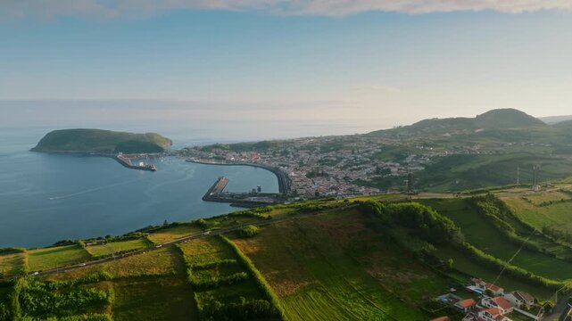 Wide aerial view of Horta city, its iconic harbor, and the volcanic Monte da Guia on Faial Island, Azores, surrounded by rolling green hills and the deep blue Atlantic Ocean.