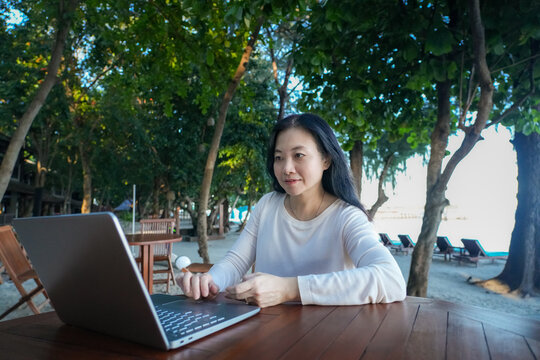 Smiling Asian woman working remotely on her laptop at an outdoor beach resort cafe, digital nomad and freelance lifestyle concept.