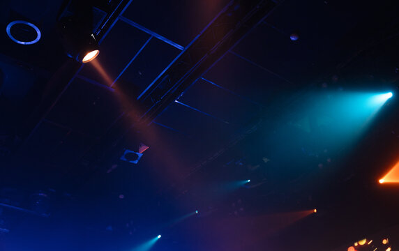 Colorful spotlights and stage lighting cut through haze above a dark concert venue ceiling, creating dramatic blue and orange beams and an atmospheric mood for live music and performance events