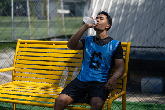 A Thai Asian soccer player drinks from a water bottle while resting on a yellow bench beside an outdoor field during a break from play