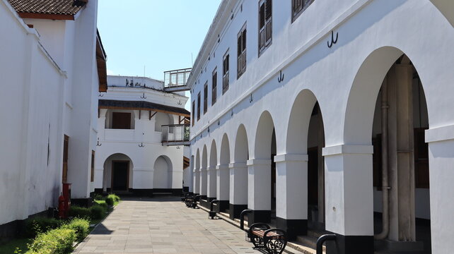 A neat, beautiful lawn featuring a black iron garden bench sits in front of an old white building with numerous arched doorways.