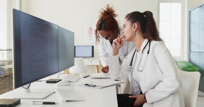 Doctors show teamwork at computer for records consultation. Two physicians review charts on dual monitors, coordinating diagnosis and updating files. Concept of digital healthcare teamwork.