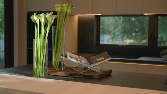 Modern kitchen with dark countertop and soft natural light, two tall glass vases hold white calla lilies adding elegance and freshness. Wooden stand with open magazine