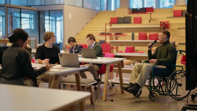 A man in a wheelchair engages his colleagues with a microphone during an active business meeting. Diverse businesspeople are attentively listening and taking notes in a modern office environment