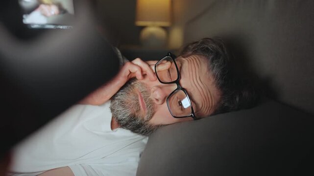 A man in eyeglasses lying in bed using smartphone late at night