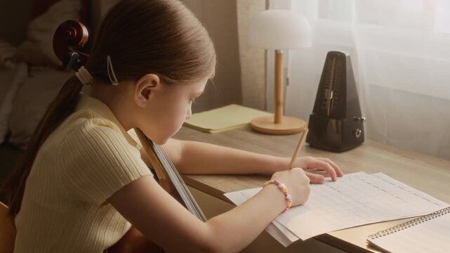 Handheld shot of child holding cello and writing notes on sheet music at desk with metronome. Footage is ideal for music education, homeschooling, and classical instrument training commercials