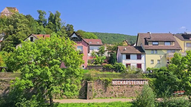 Neckarsteinach, Germany town sign on a stone wall below colorful hillside houses, lush green trees and forested hills, sunny village scene in the scenic Neckar Valley countryside.