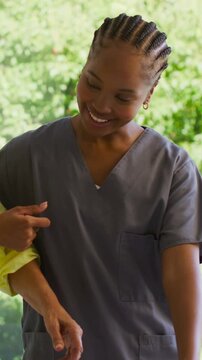 Vertical video: Smiling RN in grey scrubs guiding client hand to aid as yellow arm enters by window