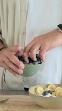 Vertical video: Lifting green bowl man in tan top adding berries then seeds into bowl for meal