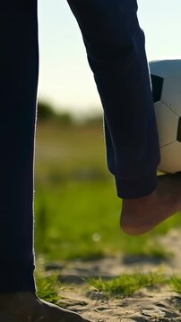 Vertical video: Balancing barefoot player lifting foot, soccer ball appearing on sandy grass, pants