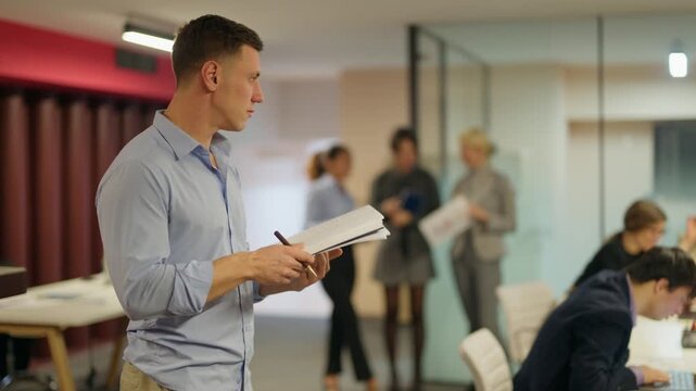 A focused businessperson writes notes in a modern office setting. His colleagues are also busy working in the open-plan workspace