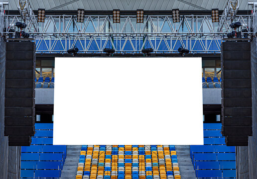 A large blank white billboard is installed on a metal stage structure with professional speakers and lighting equipment in an outdoor stadium.
