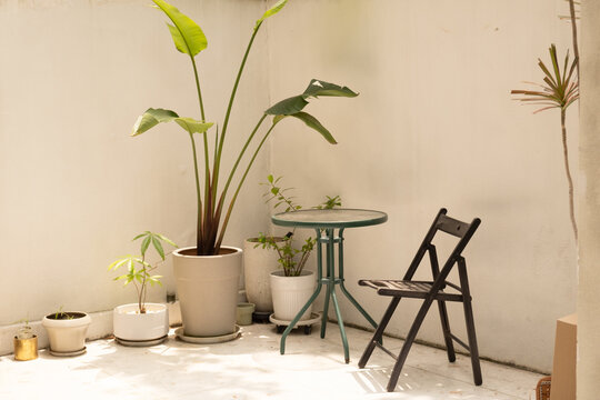 Green metal table and folding chair near potted plants in bright corner. Cozy home interior styling and relaxation space concept for modern apartment balcony.
