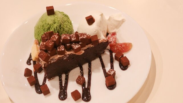 close up of a chocolate brownie cake with green tea matcha ice cream and cream on the sides. Melt chocolate suace and chocolate dice added as decoration on a white plate with copy space