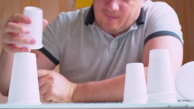 Man lining up upside-down plastic cups on a table. Blurred silhouette. Close-up, shallow DoF, natural light, cinematic bokeh. Concept of order, structure, alignment and organization.