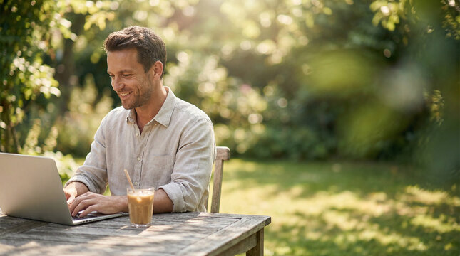 A smiling man in a linen shirt sitting at a wooden table in a green, sun-drenched garden, working on a laptop with an iced coffee. Illustrates the freedom of remote work.