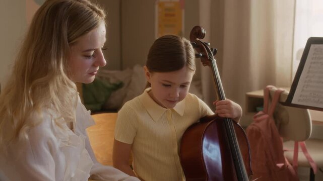 Handheld shot of female teacher explaining sheet music to Caucasian child holding cello during home music lesson. Footage ideal for music school promotions and private tutoring advertisements