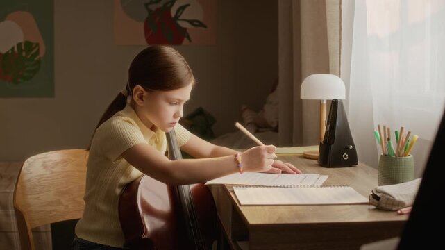 Side view of Caucasian child girl holding cello and writing in notebook at desk during music lesson. Clip is perfect for music school commercials, e-learning platforms, and educational marketing