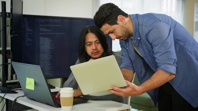 Two male software developers consulting together on laptop showing code while large monitor displays programming language in modern tech office. Diverse workplace, code review, multicultural team.