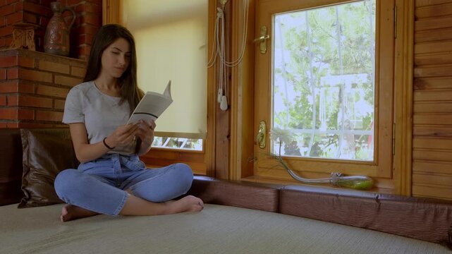 Young woman reads a book while sitting in a warm wooden cabin interior with rustic decor, soft light and calm atmosphere. Ideal for lifestyle, education, leisure, relaxation, peaceful home and cozy li