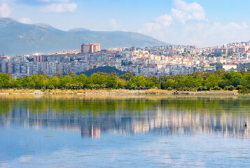 Obraz premium Scenic view of a coastal hillside city and green trees reflected beautifully on the calm water surface with majestic mountains in the background.