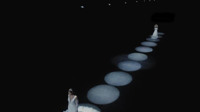 Cinematic wide shot of two elegant models in white wedding gowns standing on dark runway illuminated by circular LED ring lights at bridal fashion show, shot from elevated angle. Perfect for wedding a