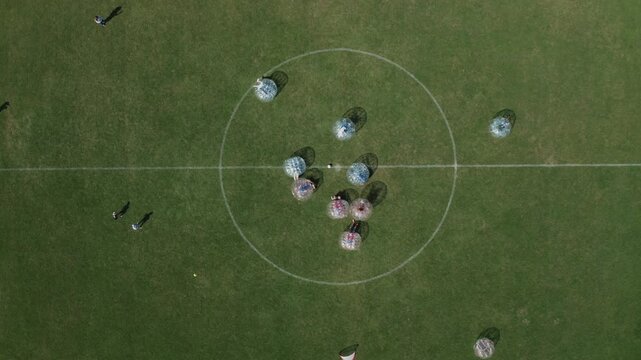 Straight-down aerial drone shot of group of people playing bubble soccer in inflatable zorb balls on green grass field. Perfect for fun, sport, entertainment, and team activity content.