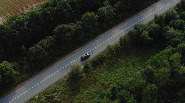 High aerial drone shot of black car driving along straight rural road between green trees and harvested wheat field. Perfect for travel, road trip, automotive, and countryside content.