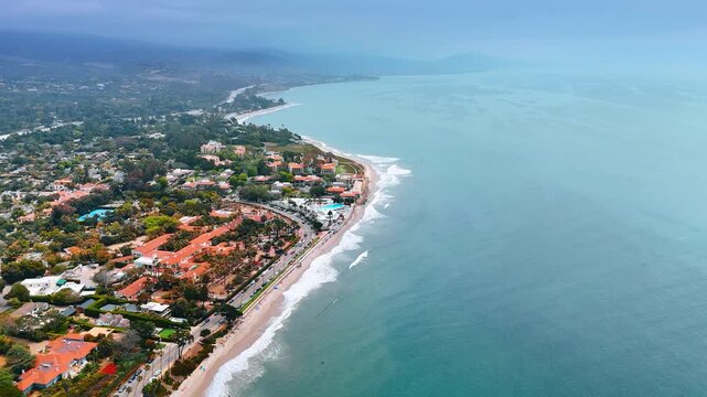 White foamy waves roll to the narrow sandy beach of the Pacific. Picturesque green scenery of modern Santa Barbara, California, USA from drone.