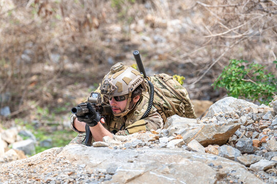 Soldier in camouflage uniform lying in prone position aiming rifle behind rocks. Tactical military warrior holding weapon during combat training outdoor