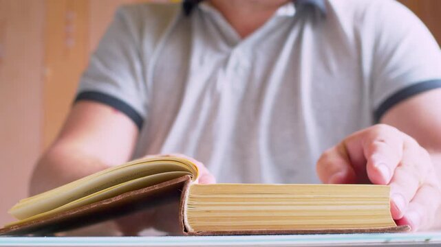 A man's hands open an old book lying on a table. A blurred silhouette of a man in a cozy modern room. Close-up, shallow depth of field, warm natural light, cinematic bokeh.