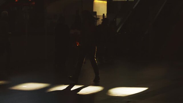 Silhouetted figures walking through a brightly lit area of a public space with shadows cast on the floor, showcasing movement and interaction in a bustling environment