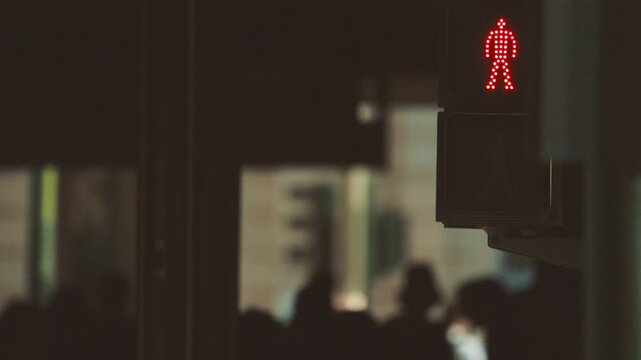 Red pedestrian traffic light signals stop for pedestrians in urban setting, with blurred figures of people waiting to cross the street in the background