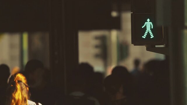 Pedestrian traffic light displaying green signal as individuals prepare to cross street, with blurred crowd and urban setting in the background, emphasizing movement and transition