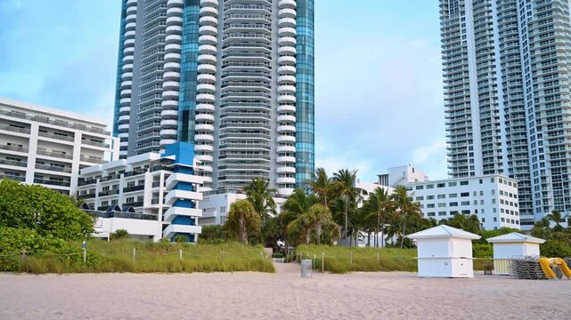 Beach huts and modern highrise residential towers in Miami, Florida. Ground perspective of white structures on sandy shore with skyscrapers under cloudy daytime sky.