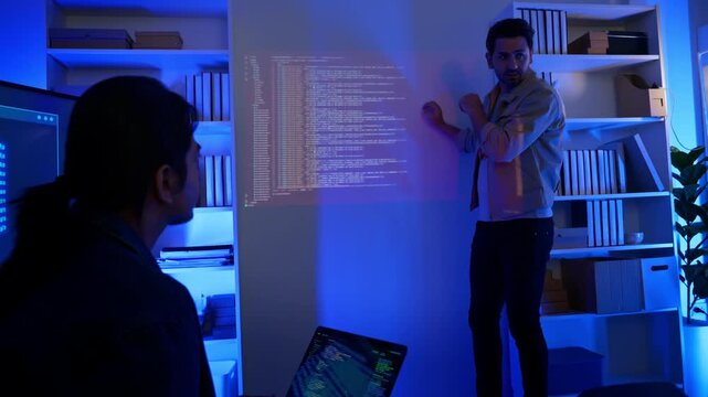 Male software developer presenting programming code projected on wall to audience in modern dark office. Tech talk, code walkthrough, bootcamp, mentorship, hackathon presentation, training session.