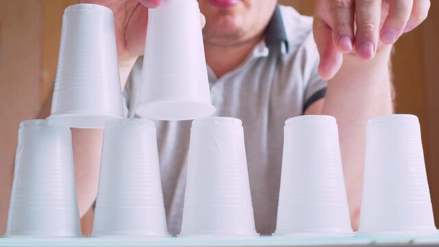 Man's hands building a pyramid with plastic cups. Blurred silhouette in a  room. Close-up, shallow depth of field, natural warm light, cinematic footage.