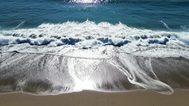 Dynamic ocean waves crash onto a sandy shoreline, creating intricate patterns of foam and water flow under a bright, sunlit sky, evoking a sense of oceanic power and serene coastal beauty