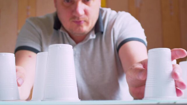 Close-up of hands stacking plastic cups into a fragile tower. Blurred male silhouette. Shallow depth of field, natural light. Metaphor for risk, business growth, stability and delicate balance.