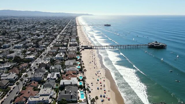 Expansive aerial perspective showcases a vibrant coastal community where golden sandy shores meet the vast expanse of the shimmering ocean, punctuated by a distant pier and the gentle rhythm