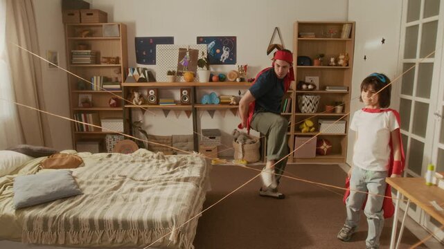 Father and young boy wearing superhero capes navigating yarn laser maze in bedroom. Video is ideal for promoting family bonding, creative indoor play, and parenting brands