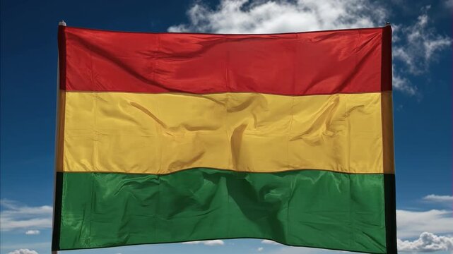 National flag of Bolivia, horizontal stripes red, yellow, green, waving proudly against clear blue sky, fluffy white clouds, symbol of nation identity, sovereignty. August 6 Bolivia's Independence Day
