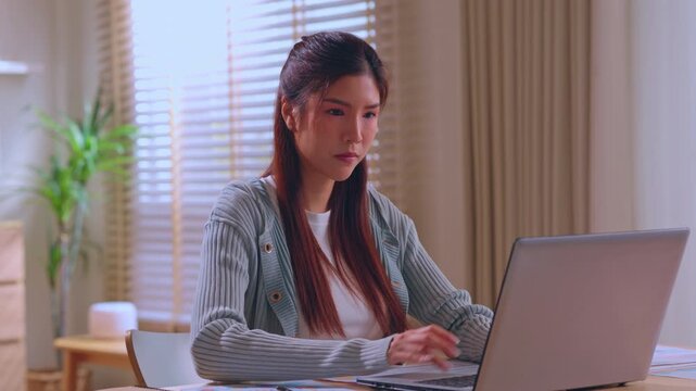 Focused woman working remote online learning on laptop computer at home office desk near window blinds, typing in cozy indoor workspace with plant and natural light