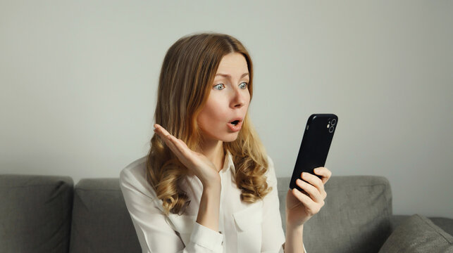Emotional surprised woman looks at phone, girl with smartphone sitting on the couch in room at home