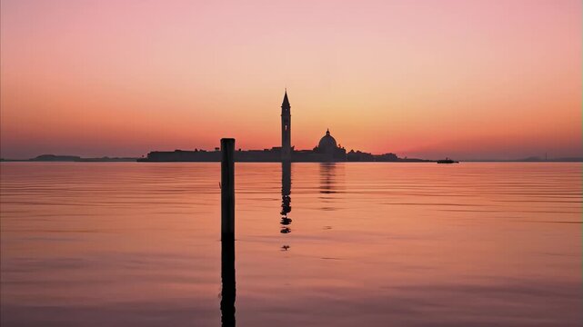 Island cityscape silhouette at sunset with prominent bell tower and dome reflection on still water and wooden pole in foreground creating a serene scene for Venice Film Festival late